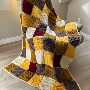 May include: A crocheted patchwork blanket in shades of yellow, brown, white, and burgundy. The blanket is made of different textured squares and is draped over a chair.
