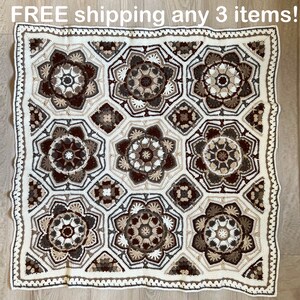 May include: A handmade crochet blanket featuring a repeating floral pattern in shades of cream, brown, and beige. The blanket has a square shape with a decorative border. The overall design is intricate and detailed, showcasing the craftsmanship of the piece.