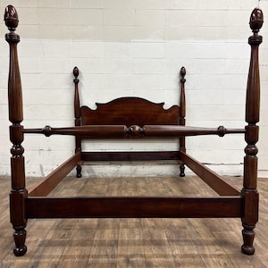 May include: A dark brown four-poster bed frame. The bed has a headboard with a curved top and four tall, turned posts. The posts are topped with finials. The bed frame is made of wood and has a classic design.