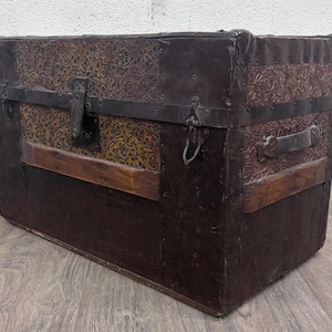 May include: A rectangular storage trunk featuring a dark brown exterior with embossed floral patterns on the upper section. The chest includes black metal corner reinforcements, horizontal metal bands, a central latch, and a side handle made of dark leather.