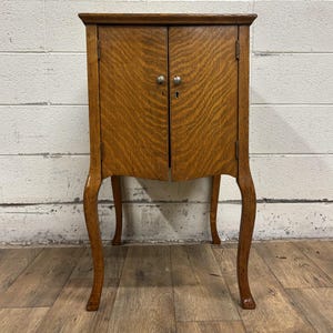 May include: A small, antique wooden cabinet with two doors and curved legs. The cabinet is made of a light brown wood with a prominent grain pattern. It has two small silver knobs and a keyhole. The legs are slender and curved, adding a touch of elegance.