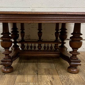 May include: A dark brown, antique-style table with an oval top and ornate, turned legs. The table features a decorative apron with small balusters. The wood has a rich, warm tone, and the table is set on a wooden floor.