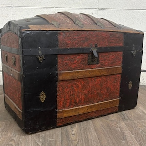 May include: A large, rectangular storage trunk with a domed lid, featuring a textured red surface with black leather-like trim, wooden slats, and metal hardware. The chest includes a central latch, decorative metal accents, and reinforced corners.