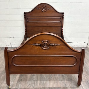 May include: A dark brown antique bed frame with a curved headboard and footboard. The headboard features a decorative carved design at the top and a rectangular panel. The footboard has a similar design. The bed frame rests on small caster wheels.