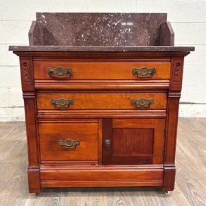 May include: A wooden washstand featuring a dark red marble top with a matching backsplash. The piece includes two full-width drawers at the top, one smaller drawer on the left, and a single cabinet door on the right, all with ornate metal hardware.