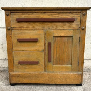 May include: A vintage wooden cabinet with a single drawer and a door. The cabinet has a light brown finish with a distressed look. The drawer and door have red handles.