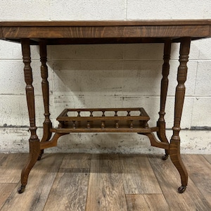 May include: A vintage wooden side table with a rectangular top and ornate, turned legs. The table features a lower shelf with a decorative railing and small caster wheels. The wood has a warm, medium-brown tone, showcasing the natural grain.
