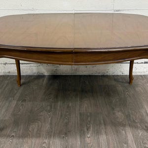 May include: An oval wooden dining table with a smooth, polished brown surface and a visible seam down the center. The table features four curved cabriole legs that extend outward from the apron, showcasing a consistent wood grain finish throughout.