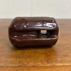 May include: A close-up of a brown ceramic insulator with a hole through the center. The insulator has a rounded rectangular shape and is likely used in electrical applications. The surface has a glossy finish.