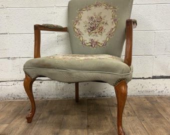 May include: An antique armchair with a light green upholstered seat and back. The back features a floral embroidery design. The chair has wooden armrests and legs with a curved design. The legs are a dark brown colour.