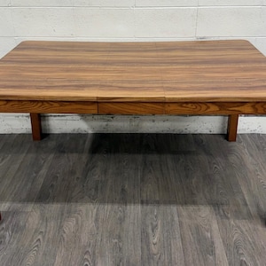 May include: A rectangular wooden dining table featuring a warm brown finish with a prominent natural wood grain pattern. The table has four straight legs and a smooth, flat surface with a visible seam indicating an extendable leaf design.