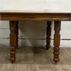 May include: A rectangular wooden table with a dark brown finish. The table has a simple design with four ornate, turned legs. The legs are supported by small caster wheels. The table is likely an antique or vintage piece.