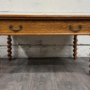 May include: A wooden coffee table with a drawer and ornate brass handles. The table has four spiral-turned legs and a warm, natural wood finish. The table is approximately 48 inches long.