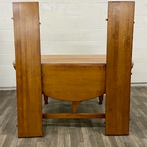 May include: A wooden drop leaf table with two leaves that are folded up and standing on end. The table is made of light brown wood and has a rounded top.