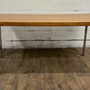 May include: A rectangular wooden table with a light brown surface and chrome legs. The table is approximately 60 inches long and 30 inches wide, with a simple, modern design. The legs are angled and provide a sturdy base.