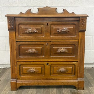 May include: A vintage wooden dresser with three drawers and ornate carvings. The dresser has a dark brown finish and a decorative top.