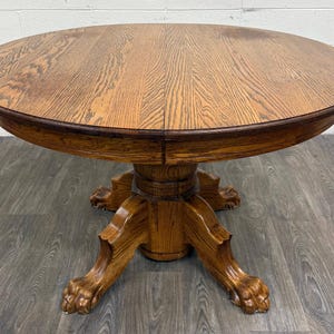 May include: A round wooden dining table with a medium brown finish and visible wood grain. The table features a central pedestal base that branches into four curved legs, each terminating in a carved lion paw foot design.
