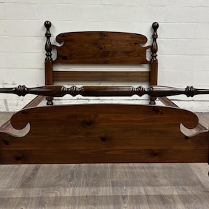 May include: A dark brown wooden bed frame featuring a headboard and footboard with curved top edges. The frame includes four tall posts topped with spherical finials and a horizontal rail connecting the front posts, showcasing a natural wood grain.