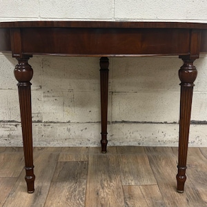 May include: A mahogany-colored, half-moon table with a rounded top and three legs. The legs are fluted and have a bulbous design near the top. The table is approximately 30 inches wide.