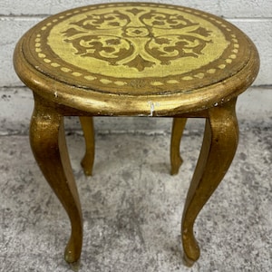 May include: A small, round, gold-painted side table with a decorative floral design on the tabletop. The table has three curved legs. The tabletop is approximately 18 inches in diameter.