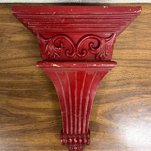 May include: A red wall shelf with an ornate design. The shelf has a triangular top with horizontal lines and a decorative scroll design. The bottom portion has vertical lines and a decorative bottom with a cone-like shape.