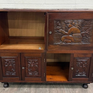 May include: A dark brown wooden cabinet featuring carved relief panels on the doors. The upper right door displays a seated figure, while the three lower doors show intricate profile carvings. The unit includes open shelving and four small feet.