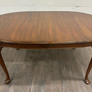 May include: An oval-shaped wooden dining table with a smooth, polished brown finish and visible wood grain. The table features four curved legs that taper toward the bottom, ending in rounded feet, providing a stable base for the tabletop surface.