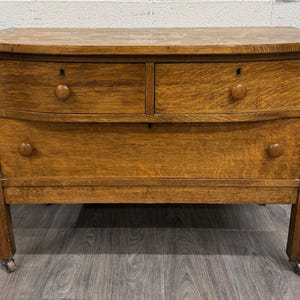 May include: A medium brown oak wood dresser featuring two smaller top drawers and one large bottom drawer. Each drawer is equipped with a single round wooden knob and a small keyhole. The furniture piece stands on four short legs with casters.