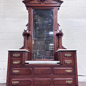 May include: A tall dark wood dresser featuring a large vertical mirror, a white marble top surface, and multiple drawers with brass hardware. The piece includes decorative side columns and a pediment top, providing a structured storage unit.