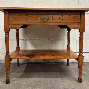 May include: A wooden side table with a drawer and a lower shelf. The table is made of a warm-toned wood and features decorative carvings and a brass drawer pull. The legs are turned and the table has a classic, antique design.