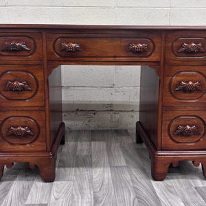 May include: A brown wooden desk featuring seven drawers, each adorned with carved floral motifs on the drawer fronts. The desk has a rectangular top, a central kneehole space, and stands on four sturdy legs with a carved base design.