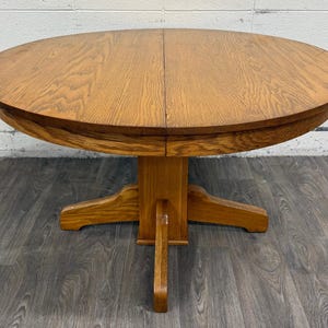 May include: A round wooden dining table with a medium brown oak finish and a visible wood grain pattern. The table features a central pedestal base with four extending feet that provide stability. The circular top has a seam down the middle.
