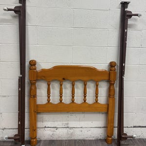 May include: A wooden bed headboard and frame set. The headboard is light brown with a curved top and vertical spindles. The frame is dark brown metal. The set is against a white brick wall.