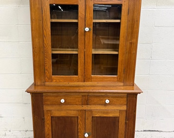May include: A tall wooden hutch featuring a natural brown finish with visible wood grain. The upper section has two glass-paneled doors with white knobs, while the lower section includes two drawers and two solid doors, all equipped with white knobs.