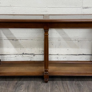 May include: A rectangular wooden console table featuring a dark brown finish, a smooth top surface with two inset glass panels, and a lower shelf with a woven cane texture. The table is supported by four vertical legs with decorative detailing.