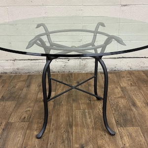 May include: A round glass-top table with a black metal base. The table has a decorative, curved design on the legs and a circular support structure. The glass top is clear, and the table is set on a wooden floor.