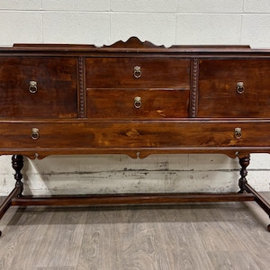 May include: A dark brown wooden sideboard with ornate detailing. It features two cabinet doors, two drawers, and a long drawer, all with brass ring pulls. The sideboard has decorative legs and a scalloped apron.