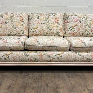 May include: A three-cushion sofa with a light wood frame and floral patterned cushions. The cushions and base feature a cream background with pink, yellow, and green floral designs. The sofa has a curved armrest design.