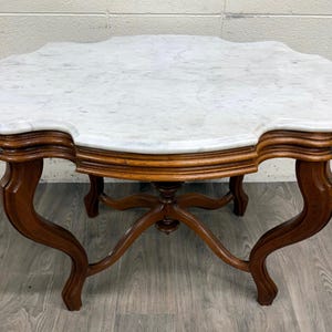 May include: An antique coffee table with a white marble top and a scalloped edge. The table has a dark wood base with curved legs and a detailed pedestal. A classic piece of furniture.