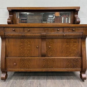 May include: A brown oak sideboard featuring a rectangular mirror attached to the top, four small upper drawers, two central cabinet doors with round wooden knobs, and one large bottom drawer spanning the width of the base with curved side supports.