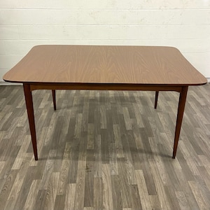Puede incluir: Una mesa de comedor de madera marrón con una tapa rectangular y cuatro patas. La mesa está sobre un suelo de madera.