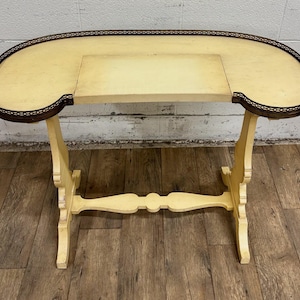 May include: An antique, cream-colored vanity table with an oval top and a rectangular inset. The table has a dark brown trim around the edge and is supported by four legs connected by a decorative stretcher. The table is made of wood.