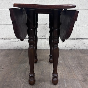 May include: A dark brown wooden drop-leaf table featuring a smooth rectangular top with two hinged side leaves that hang vertically. The table is supported by four turned legs with a rounded, tapered design and a polished wood finish.