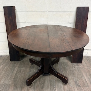 May include: A round, dark brown wooden dining table with a visible wood grain pattern. The table features a central pedestal base with four feet, each equipped with small metal casters. Two matching rectangular wooden leaves stand upright behind.