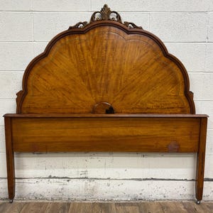 May include: A wooden headboard with a scalloped top and ornate carved details. The headboard has a sunburst pattern and is made of a warm-toned wood. The headboard is supported by legs with small wheels.