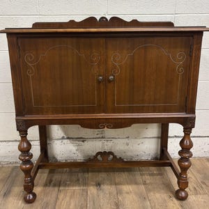 May include: A dark brown wooden cabinet with two doors and decorative silver-colored accents. The cabinet sits on a stand with ornate, turned legs and a decorative apron. The cabinet has a curved top and small, round door handles.