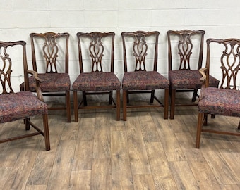 May include: Set of six antique wooden chairs with ornate backs and patterned upholstered seats. The chairs are a dark brown color and have a classic design. Two chairs have armrests. The chairs are arranged in a row.