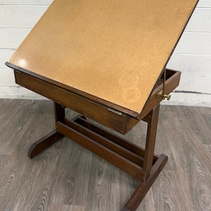 May include: A wooden drafting table with a large, light brown, angled work surface and a dark brown frame. The table features a hinged top, a storage compartment underneath, and a brass-colored metal adjustment mechanism on the side.
