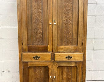 May include: A tall, wooden cabinet with a warm brown finish. The cabinet features two upper doors with carved spiral designs, two drawers, and two lower doors. It has white knobs and brass drawer pulls.