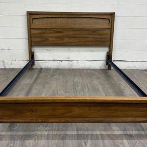 May include: A wooden bed frame featuring a tall headboard with a rectangular inset panel and a matching low footboard. The frame is constructed from medium-toned wood with visible grain patterns and includes black metal side rails for support.